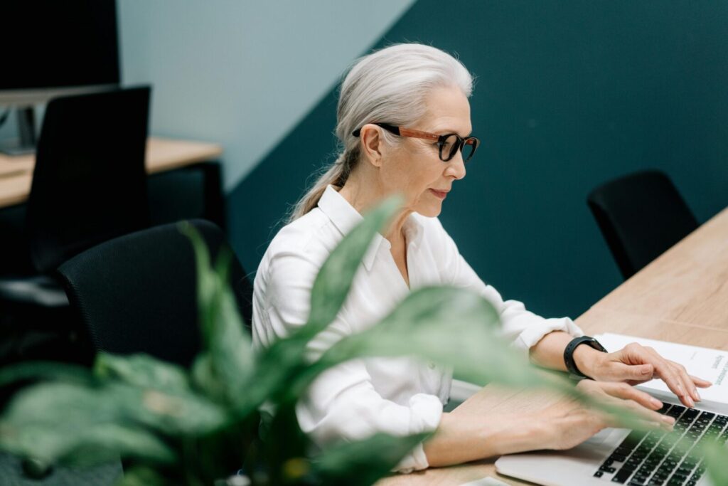 Confident senior woman with glasses working on a laptop in a modern office setting.