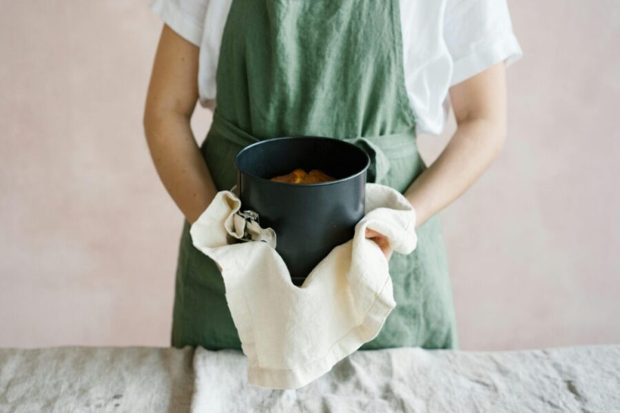 A person in a green apron holds a round baking pan with a cake inside, using a towel.