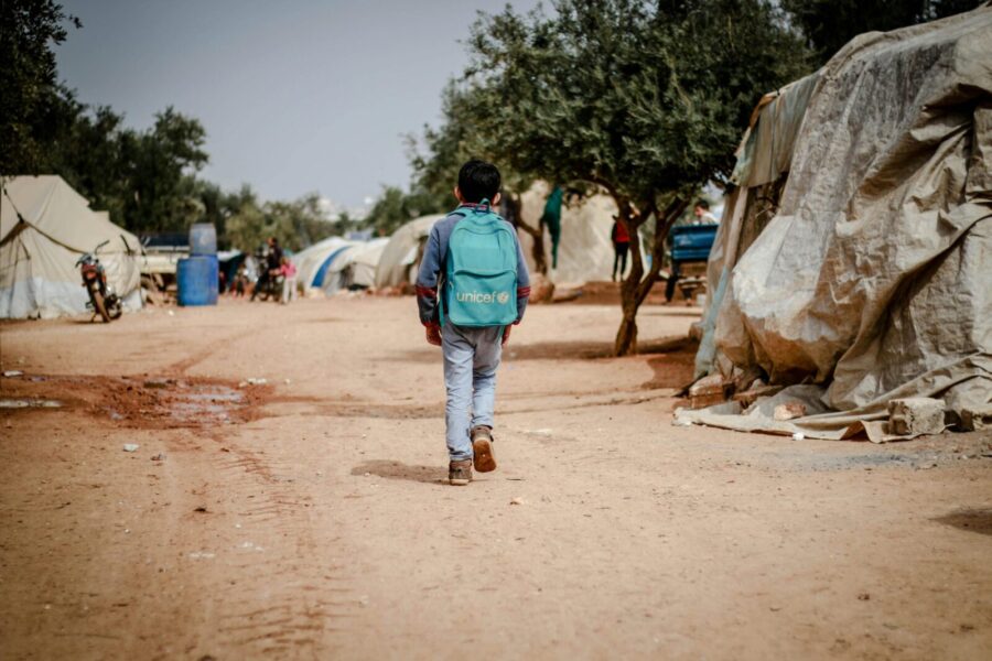 A young boy walks through a refugee camp in Idlib, Syria, wearing a backpack.