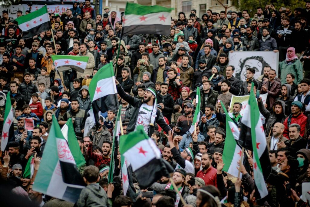 A large crowd gathers in a city square waving Syrian flags during a protest.