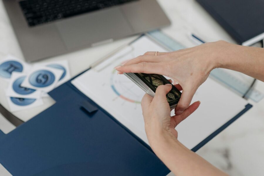 A woman shuffling tarot cards on a desk with a horoscope chart and a laptop.
