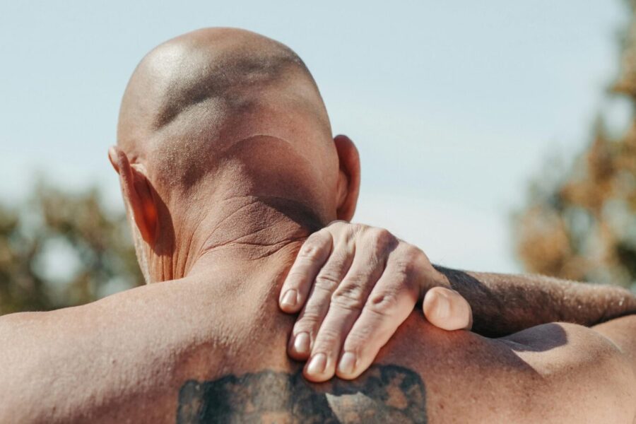 A bald man with a large tattoo massages his aching back outdoors, under clear skies.