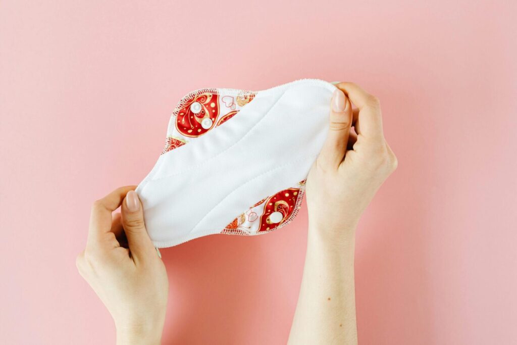 Close-up of hands holding a cloth menstrual pad with colorful design on a pink background.