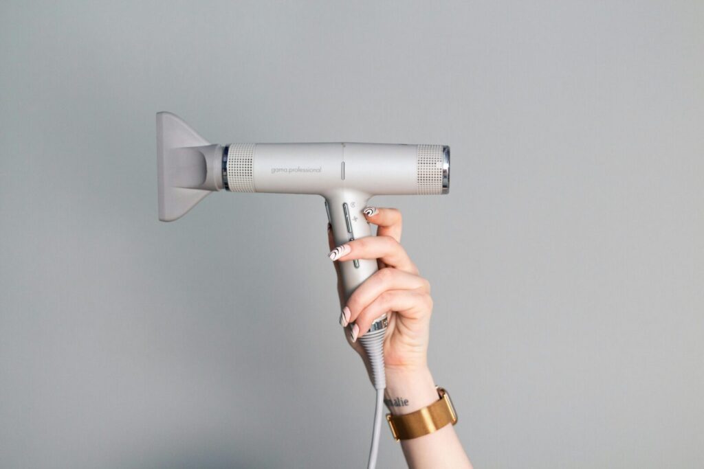 A close-up of a hand with manicured nails holding a sleek hair dryer against a neutral background.