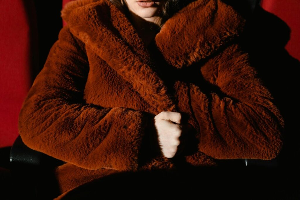 Close-up of a person warmly wrapped in a plush brown fur coat, seated indoors.