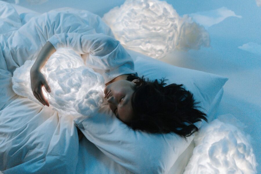 Woman in white hugging glowing cloud pillows in serene, dreamlike setting.