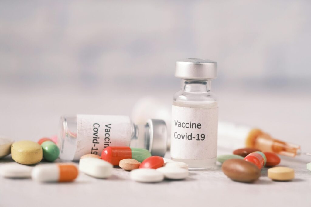 Close-up image of Covid-19 vaccine vials with assorted pills and a syringe on a light surface.