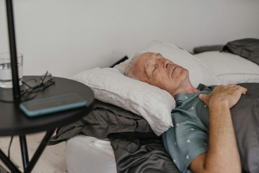 A senior man sleeps peacefully on his bed with hand on chest in a cozy bedroom setting.