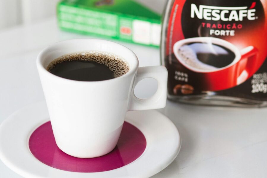 A modern white mug with black coffee sits on a pink saucer, Nescafé jar in the background.