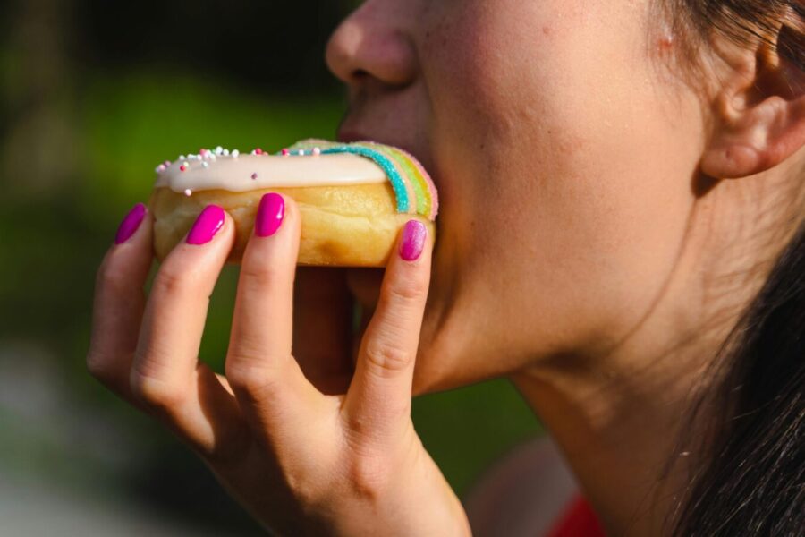 A woman with manicured nails takes a bite of a colorful sprinkled doughnut.