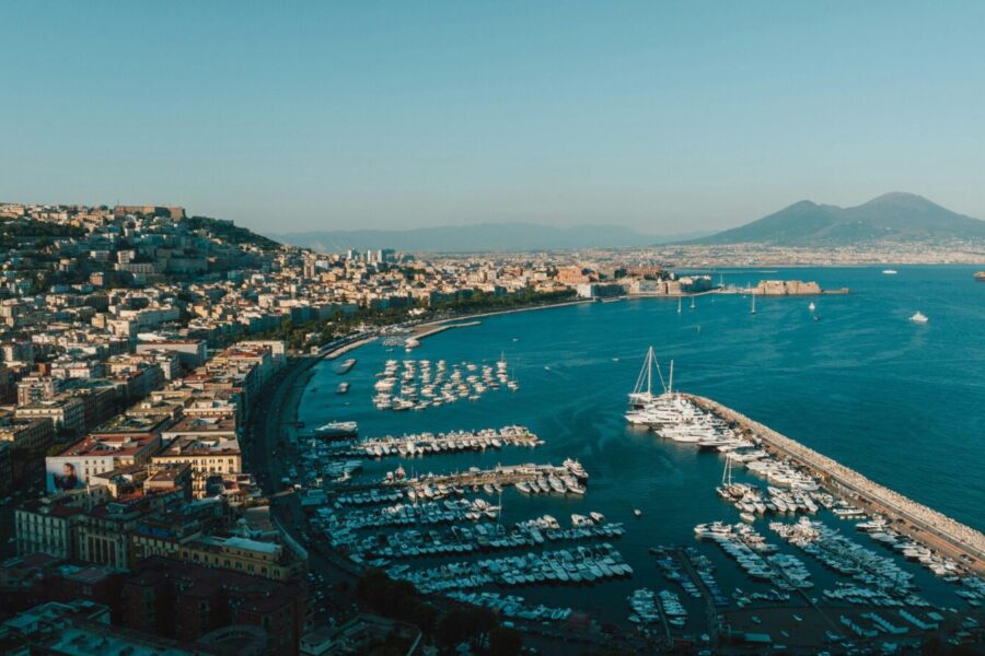 Aerial view of Naples harbor with boats and city skyline under clear blue sky.