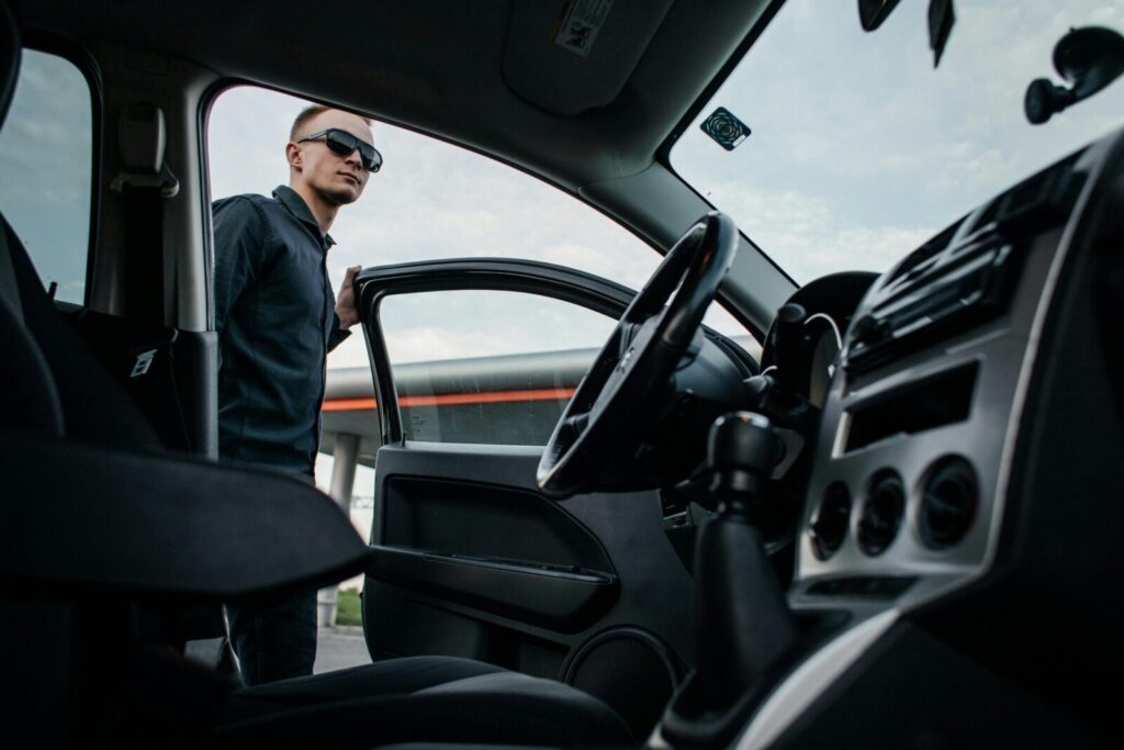 A man in sunglasses stands by an open car door, viewed from the interior.