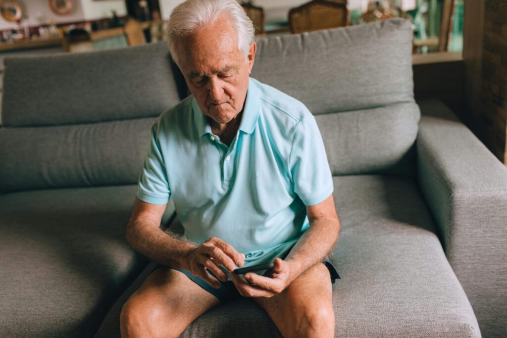 Elderly man sitting on a sofa inside a cozy living room, using a smartphone.