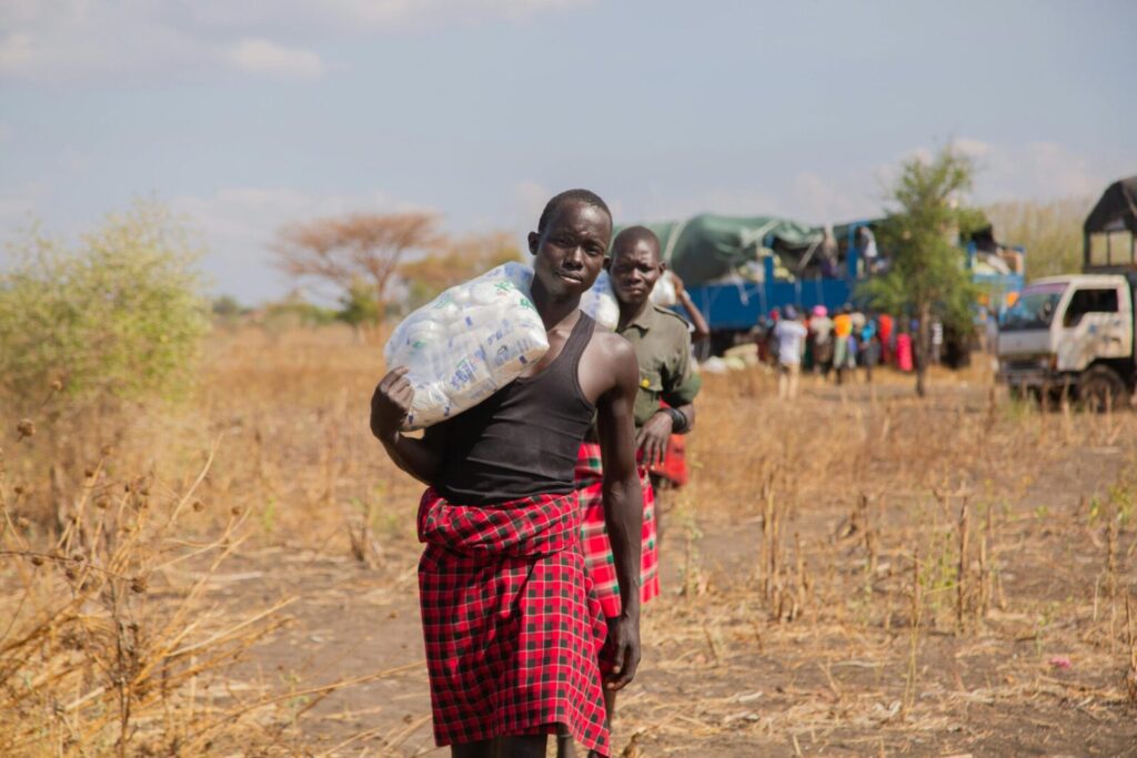 Two men in traditional attire carry supplies in an African savannah setting.