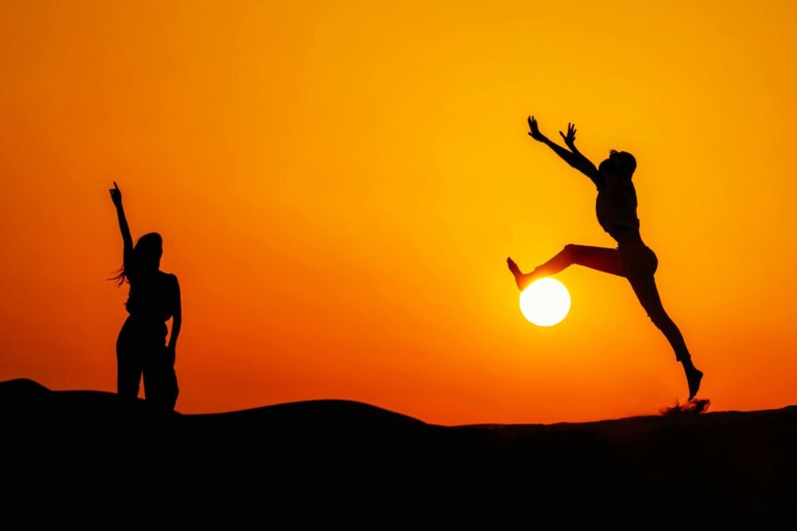 Two silhouetted people jumping joyfully during a vibrant sunset in Dubai's desert.