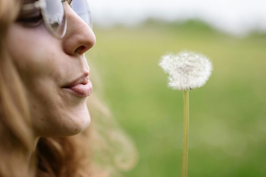 A woman in glasses blows a dandelion in a lush green field. Capturing a serene, natural moment.