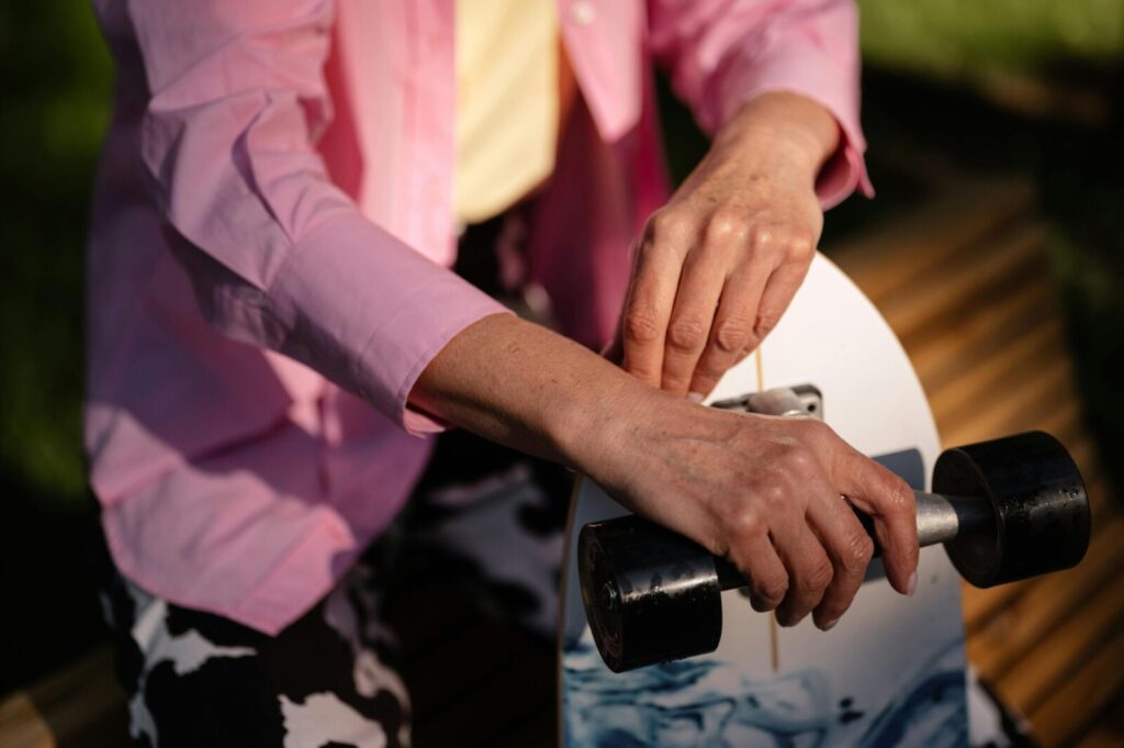 Hands holding a skateboard with pink sleeves, outdoors in natural lighting.