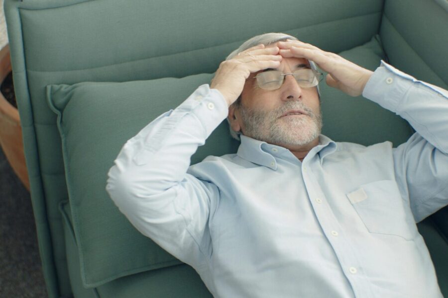 A senior man with eyeglasses relaxing on a sofa, looking stressed and tired.